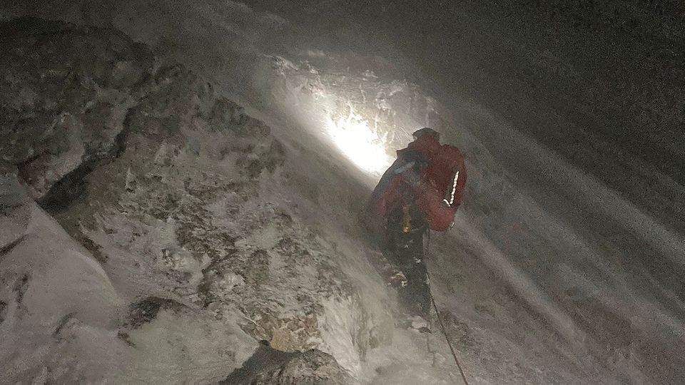 Im dunkeln steht ein Mensch in roter Kleidung an einem verschneiten Berghang. Eine Stirnlampe erhellt das Schneetreiben