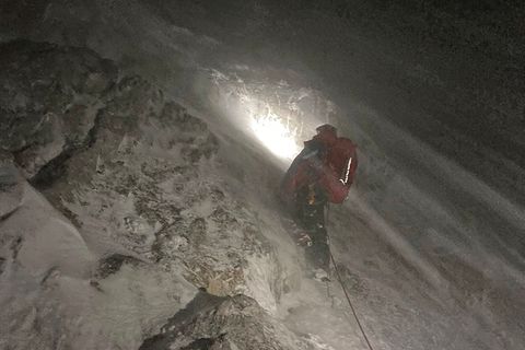 Im dunkeln steht ein Mensch in roter Kleidung an einem verschneiten Berghang. Eine Stirnlampe erhellt das Schneetreiben