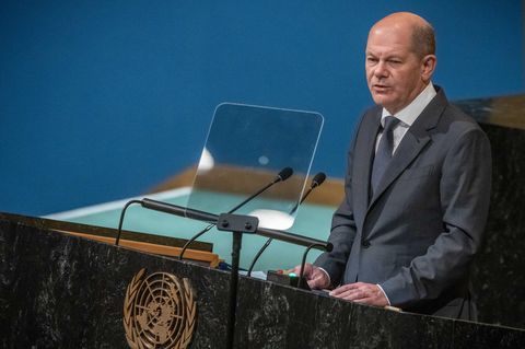 An einem schwarzen Rednerpult mir goldenem Weltkarten-Wappen auf der Vorderseite steht ein weißer Mann im Anzug