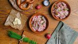 Bayerischer Wiesn Wurstsalat   Der Wurstsalat ist in Bayern ein typisches Gericht für den Biergarten.      Zutaten für 2 Personen  Vorbereitungszeit 15 Minuten  Zubereitungszeit 10 Minuten      100 ml Weißweinessig  3-4 Prisen Salz  4 Prisen Pfeffer  1 EL Zucker  1 TL mittelscharfer Senf  3 kleine, rote Zwiebeln  300 g Leberkäse (Neuburger) dünne Scheiben, 0,9 auf der Aufschnittmaschine  150 g Cornichons aus dem Glas  4 EL Sonnenblumenöl  1/2 Bund Schnittlauch      Beilagen:  2-4 frische Brezen  1 Bund Radieserl  80 g Butter      Zubereiten:  Essig in einer Schüssel mit Salz, Pfeffer, Zucker und Senf verrühren.  Die Zwiebeln halbieren und schälen.  Entweder dünne, feine Zwiebelstreifen oder feine Würfel schneiden und zum Essig geben.  Die Leberkäse-Scheiben exakt übereinander legen und zuerst 2,5 cm breite Streifen, dann 2,5 cm breite Rauten schneiden.  Die Leberkäse-Rauten auf dem Schneidebrett auseinander zupfen und dann in die Marinade legen.  Die Cornichons auf ein Sieb gießen, abtropfen lassen, anschließend der Länge nach halbieren oder in Scheiben schneiden.  Die Cornichons zum Leberkäse geben und gut vermischen. Den Salat mindestens 20 Minuten marinieren lassen.  Kurz vor dem Servieren nochmals abschmecken, nach Geschmack mit Öl ergänzen und anrichten.  Fein geschnittene Schnittlauchröllchen auf den Wurstsalat streuen und mit Brezen, Butter und Radieschen servieren.         Tipp von Koch Thomas Sixt:  Die Reihenfolge der Zubereitung ist für diesen Salat wichtig. Die rohen Zwiebeln sind, eingelegt in Essigmarinade länger haltbar. Den Salat können Sie im Kühlschrank gut einen Tag durchziehen lassen. Schnittlauch gebe ich stets frisch auf den angerichteten Salat, das Aroma ist so einfach besser.