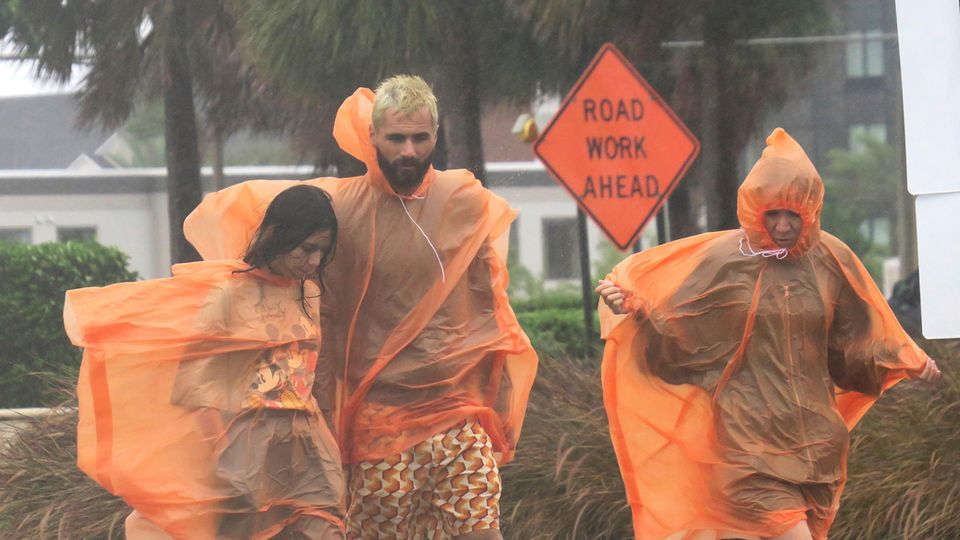 Ein Mann und zwei Frauen gehen in orangen Regencapes über eine Straße im US-Bundesstaat Florida