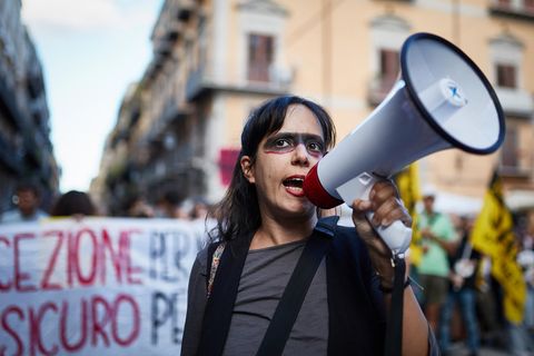 Eine Demonstrantin mit einem Megafon in einer Straße Italiens, im Hintergrund ein Plakat