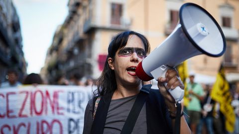 Eine Demonstrantin mit einem Megafon in einer Straße Italiens, im Hintergrund ein Plakat