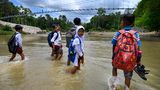 Kuta Cot Glie, Indonesien. Mit Schulranzen auf dem Rücken und Schuhen in den Händen überquert eine Gruppe von Jungen und Mädchen einen knietiefen Fluss. Im Hintergrund sieht man die Bauarbeiten an einer Hängebrücke. Bis diese fertig ist, führt der Schulweg für die Kinder aus dem Dorf Siron durchs Wasser.