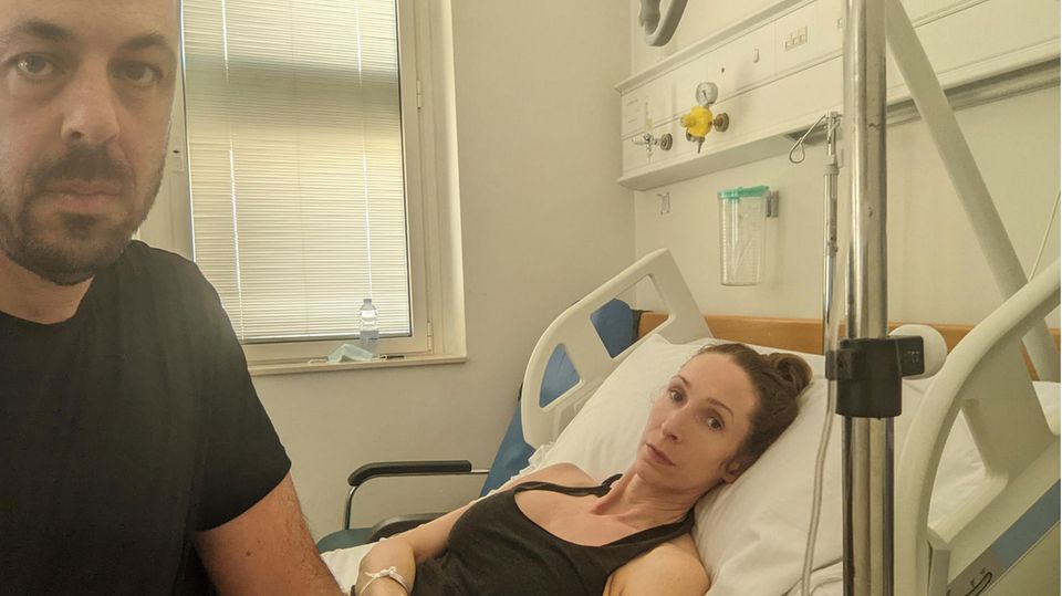 Eine Frau liegt in einem Krankenhausbett in Malta, neben ihr sitzt ihr Partner, beide schauen in die Kamera