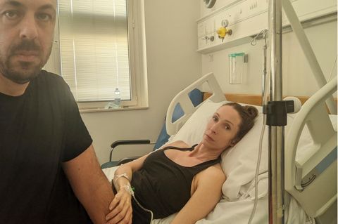 Eine Frau liegt in einem Krankenhausbett in Malta, neben ihr sitzt ihr Partner, beide schauen in die Kamera