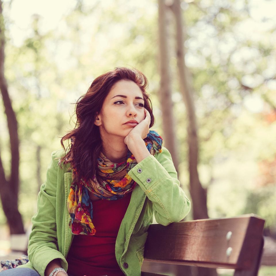 Eine Frau sitzt alleine auf einer Parkbank und guckt traurig in die Ferne.