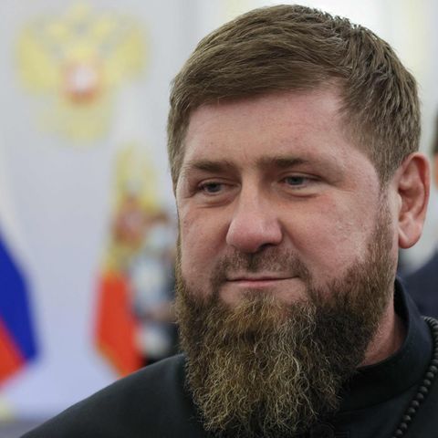 Ein weißer Mann mit dunkelblondem Haar und Vollbart lächelt leicht. Hinter ihm ein mit russischen Flaggen geschmückter Saal
