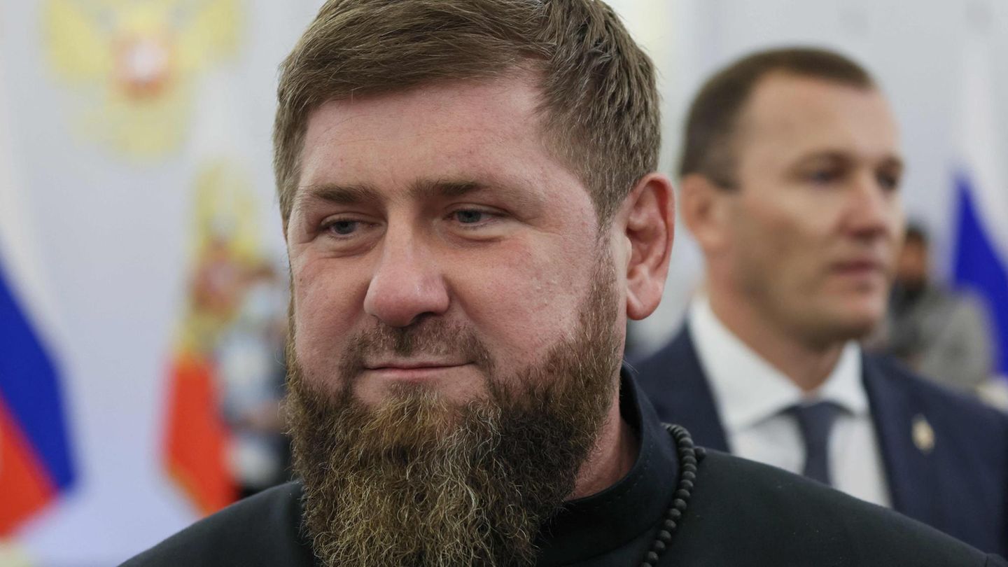 Ein weißer Mann mit dunkelblondem Haar und Vollbart lächelt leicht. Hinter ihm ein mit russischen Flaggen geschmückter Saal