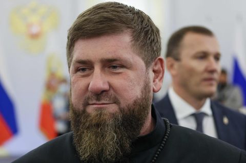 Ein weißer Mann mit dunkelblondem Haar und Vollbart lächelt leicht. Hinter ihm ein mit russischen Flaggen geschmückter Saal