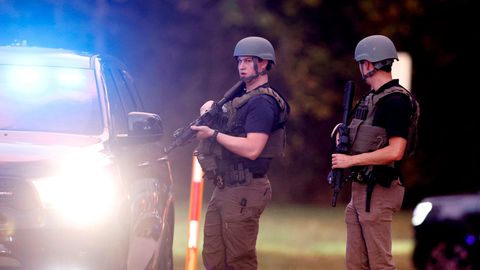 Zwei Polizisten in Schutzwesten und mit Helmen sowie Gewehren stehen neben einem Polizeiwagen mit eingeschaltetem Blaulicht