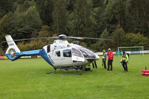 Ein silber-blau lackierter Polizeihubschrauber steht auf einer Wiese vor einem Tannenwald. Um ihn herum stehen vier Menschen