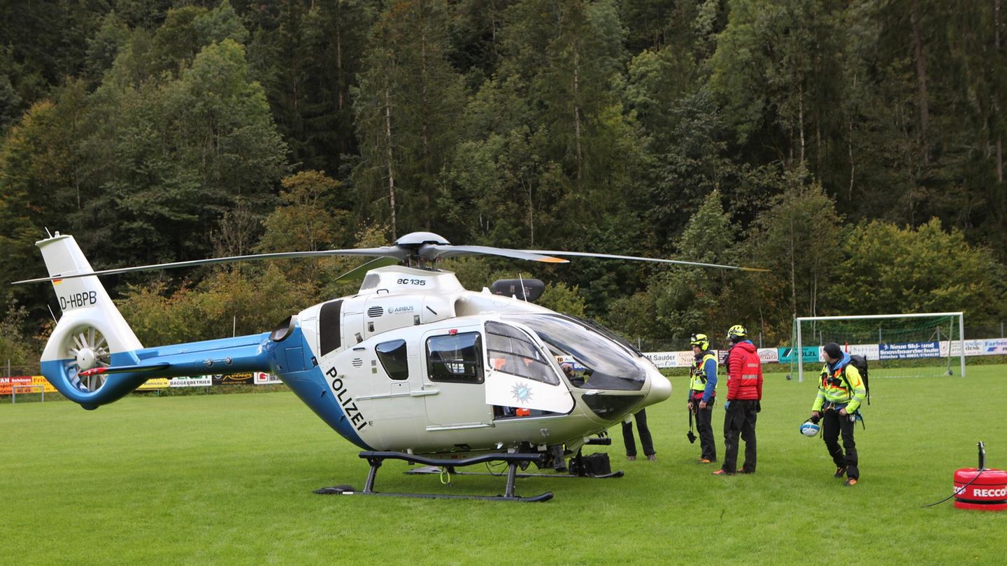 Ein silber-blau lackierter Polizeihubschrauber steht auf einer Wiese vor einem Tannenwald. Um ihn herum stehen vier Menschen