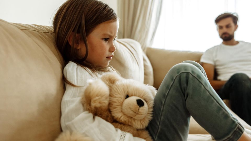 Ein Kind sitzt mit seinem Teddy traurig guckend auf dem Sofa.
