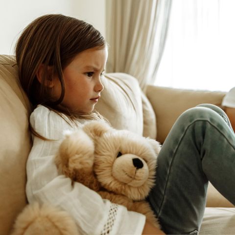 Ein Kind sitzt mit seinem Teddy traurig guckend auf dem Sofa.