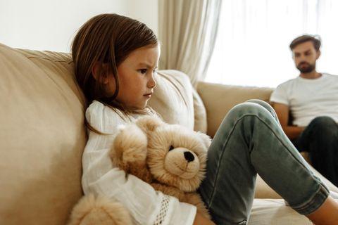 Ein Kind sitzt mit seinem Teddy traurig guckend auf dem Sofa.