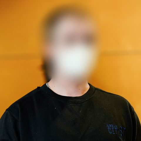 Vor einer Holzwand sitzt ein junger Mann in dunkelblauem Sweatshirt und mit weißer FFP2-Maske. Sein Gesicht ist unkenntlich