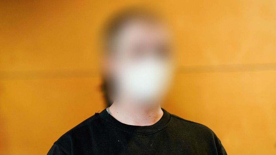 Vor einer Holzwand sitzt ein junger Mann in dunkelblauem Sweatshirt und mit weißer FFP2-Maske. Sein Gesicht ist unkenntlich