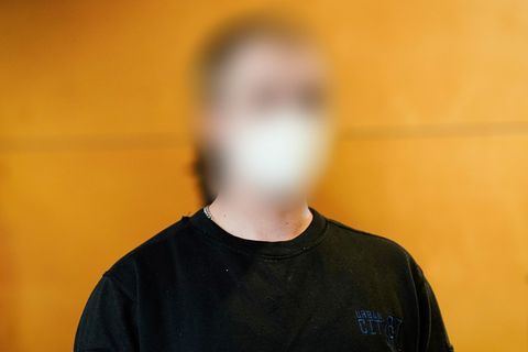 Vor einer Holzwand sitzt ein junger Mann in dunkelblauem Sweatshirt und mit weißer FFP2-Maske. Sein Gesicht ist unkenntlich