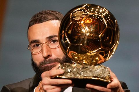 Ein weißer Mann mit braunem Haar und Vollbart trägt Brille und Anzug und hält einen goldenen Fußball in seinen Händen