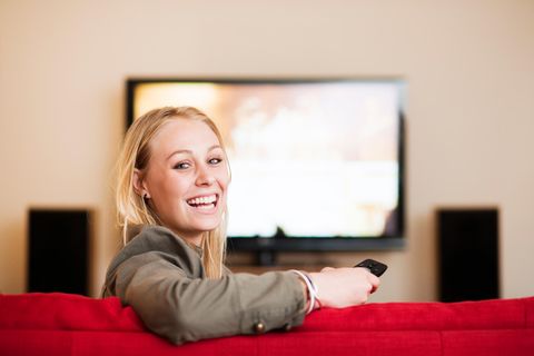 Eine Frau sitzt vorm Fernseher und schaut freudig in die Kamera