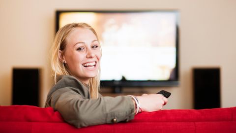 Eine Frau sitzt vorm Fernseher und schaut freudig in die Kamera