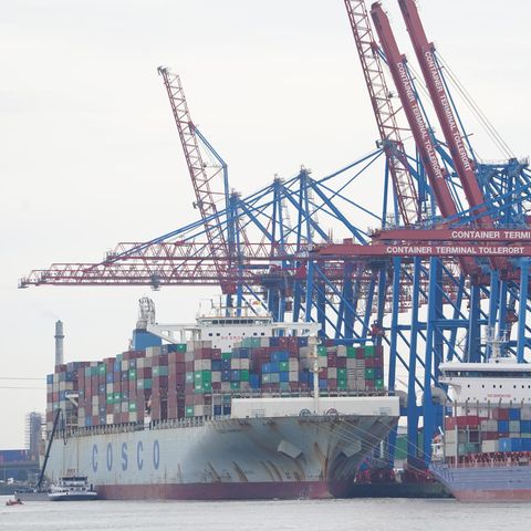 Ein graues Containerschiff mit "Cosco"-Schriftzug auf dem Rumpf liegt unter blau-roten Kränen am Terminal Tollerort
