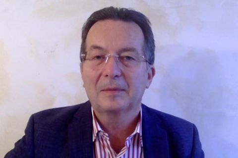 Ein weißer Mann mit dunkelgrauem Haar und randloser Brille sitzt in Jacket und rot -weiß gestreiftem Hemd vor einer hellen Wand