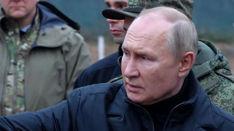 Wladimir Putin zeigt mit einer Brille in der Hand auf etwas beim Besuch eines militärischen Trainingszentrums