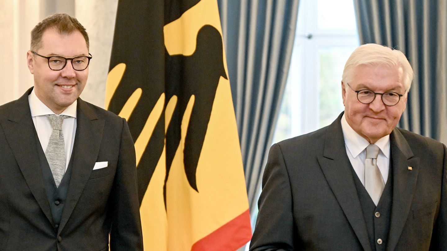 Zwei weiße Männer in Fracks stehen vor einer Flagge: schwarzer Bundesadler auf gelbem Grund mit dünnem rotem Rand