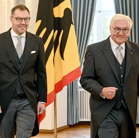 Zwei weiße Männer in Fracks stehen vor einer Flagge: schwarzer Bundesadler auf gelbem Grund mit dünnem rotem Rand