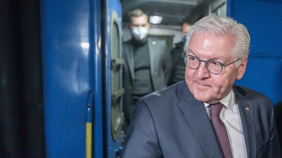 Ein weißer Mann mit weißem Seitenscheitel steigt in Anzug und Krawatte aus einem blauen Eisenbahnwaggon