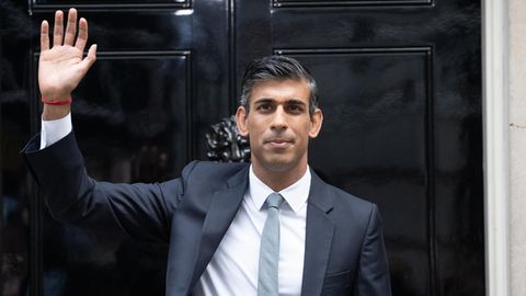 Ein indischstämmiger Mann steht in Anzug und Krawatte vor einer schwarz lackierten Haustür mit einer 10