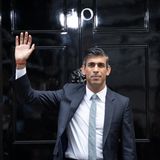 Ein indischstämmiger Mann steht in Anzug und Krawatte vor einer schwarz lackierten Haustür mit einer 10