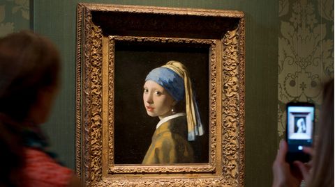 Besucher betrachten und fotografieren ein Ölgemälde, das ein weißes Mädchen mit blau-gelbem Kopftuch und Perlenohrring zeigt