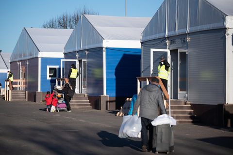 Menschen in Winterkleidung gehen mit Rollkoffern und weißen Plastiktüten an Hallen mit Planen-Dächern vorbei