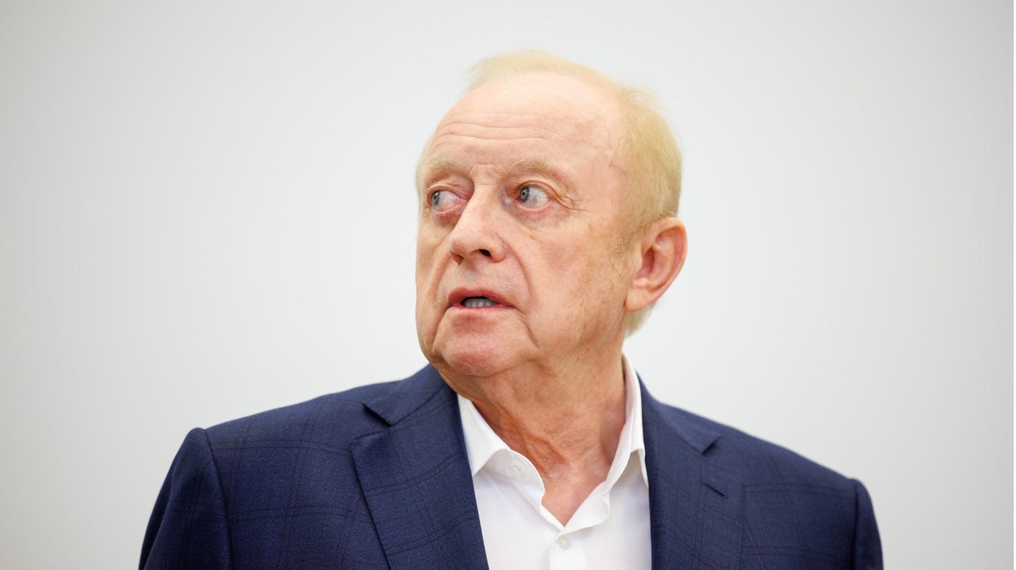 En alter weißer Mann mit schütterem blonden Seitenscheitel steht in blauem Jackett und weißem Hemd in einem Gerichtssaal