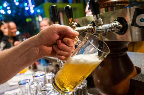 Die linke Hand eines weißen Mannes hält ein Bierglas schräg unter einen Zapfhahn, aus dem Bier fließt