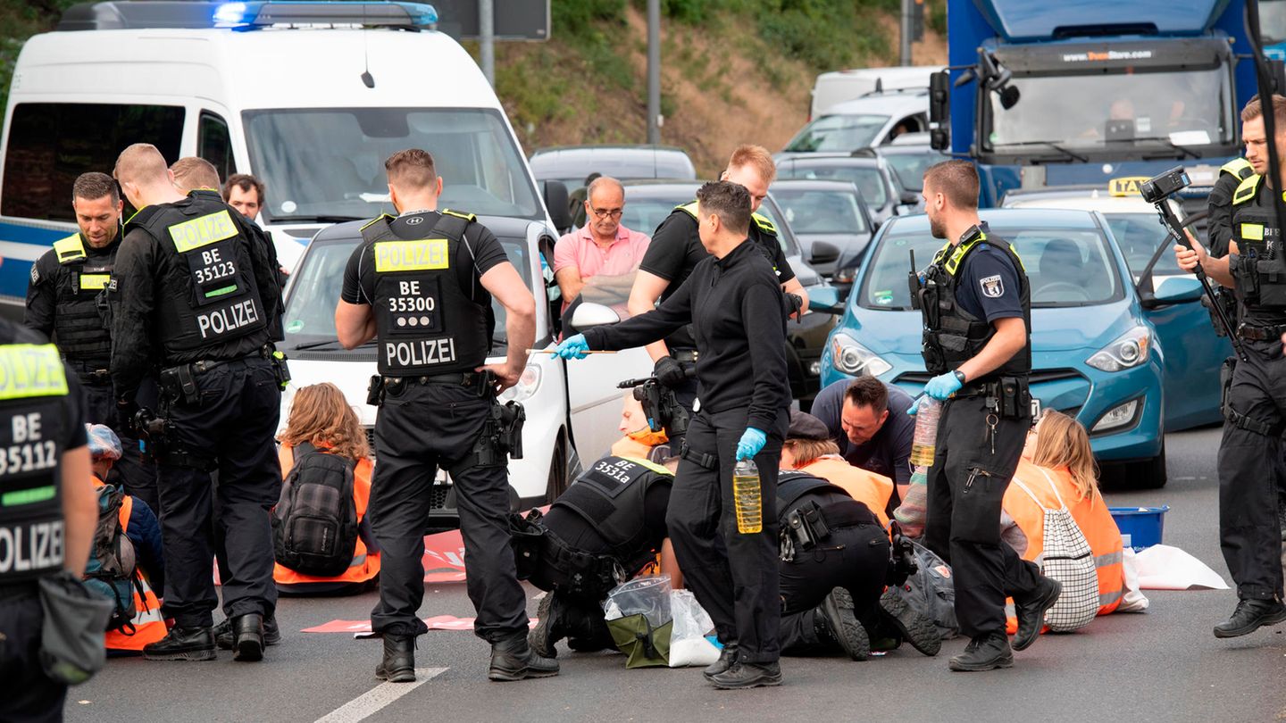 Junge Menschen in orangen Warnwesten sitzen zwischen Autos auf einer mehrspurigen Straße. Polizisten stehen hinter ihnen