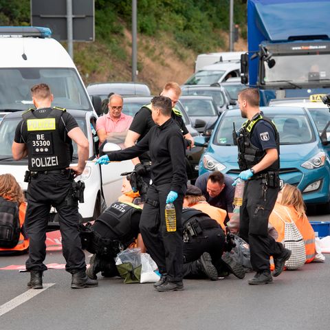 Junge Menschen in orangen Warnwesten sitzen zwischen Autos auf einer mehrspurigen Straße. Polizisten stehen hinter ihnen