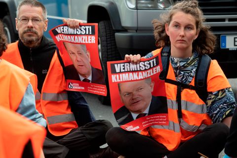 Ein Mann und eine Frau in orangen Warnwesten sitzen vor einem Polizeiauto auf einer Straße und halten Protestplakate hoch