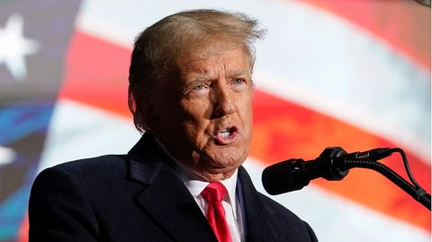 Ein alter weißer Mann mit dünnem graublonden Seitenscheitel steht in Anzug und roter Krawatte vor einer US-Flagge und spricht