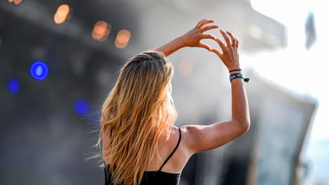 Eine junge, blonde Frau formt vor einer Festivalbühne mit Daumen und Zeigefinger beider Hände ein Herz