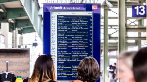 Menschen stehen vor einer Anzeigetafel mit Deutsche Bahn-Fahrplan
