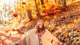 Eine Frau steht lachend unter herabfallendem Herbstlaub.