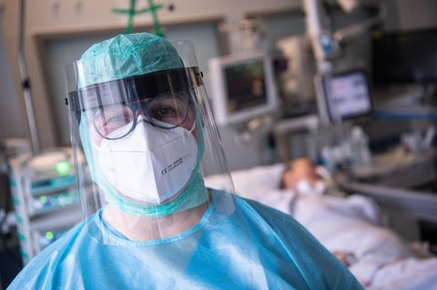 Ein Mensch in blauer und grüner medizinischer Schutzkleidung sowie Gesichtsvisier und FFP-Maske steht vor einem Intensivbett