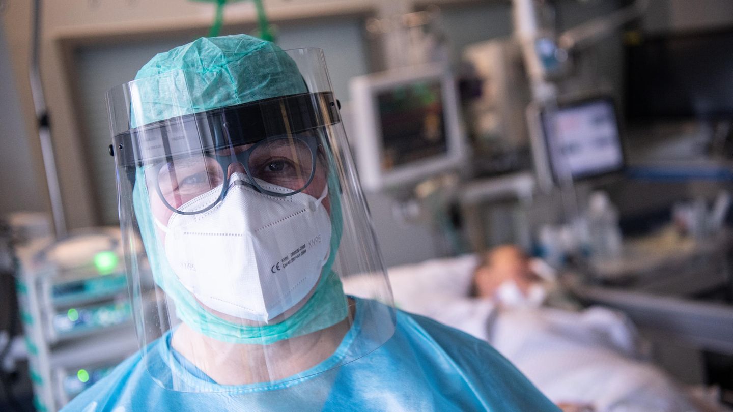 Ein Mensch in blauer und grüner medizinischer Schutzkleidung sowie Gesichtsvisier und FFP-Maske steht vor einem Intensivbett