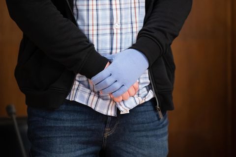 Ein weißer Mann in Karohemd, Jeans und Strickjacke steht in einem Gerichtssaal und umfasst mit einer Hand das andere Handgelenk