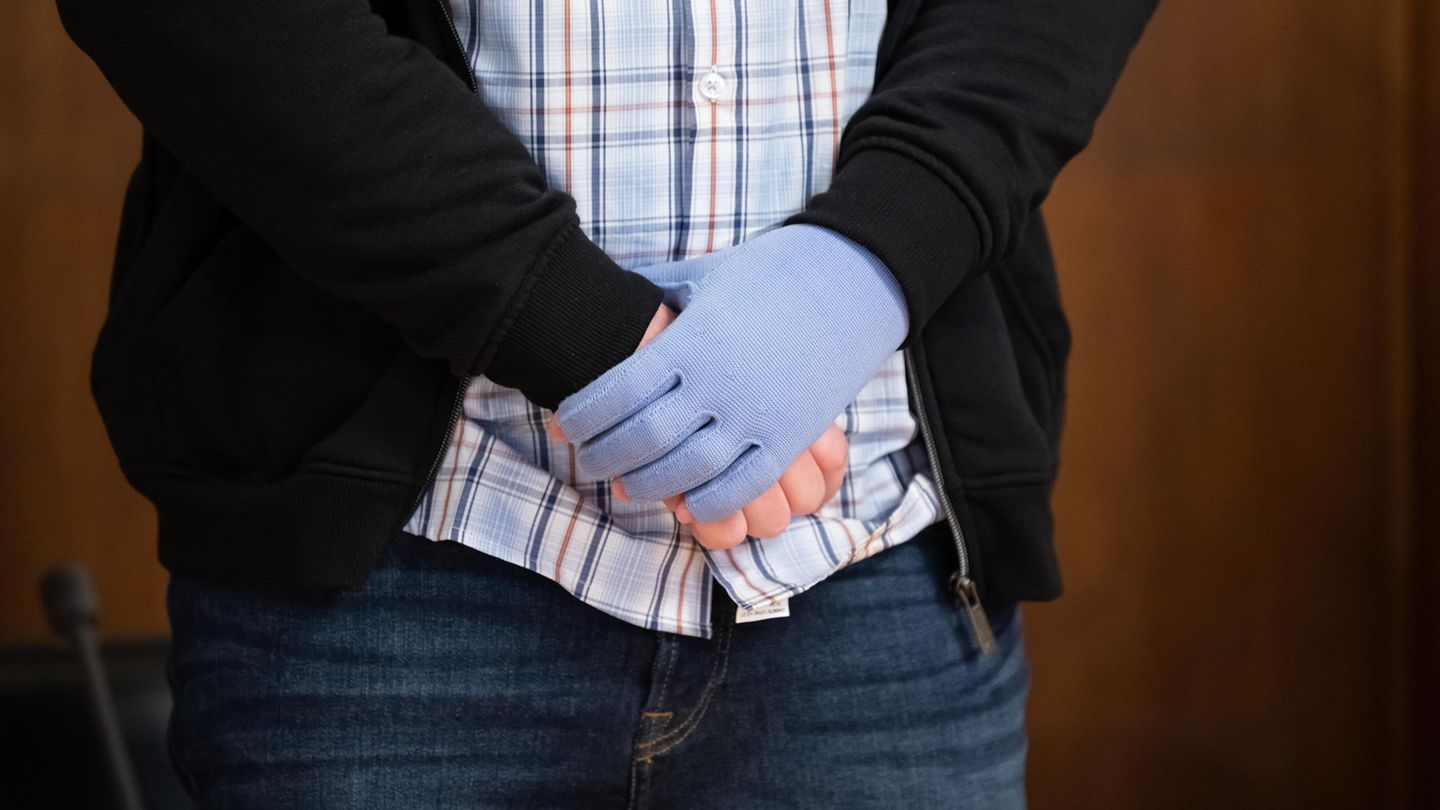 Ein weißer Mann in Karohemd, Jeans und Strickjacke steht in einem Gerichtssaal und umfasst mit einer Hand das andere Handgelenk