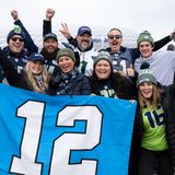 Männer und Frauen in Football-Trikots und warmer Kleidung jubeln hinter einer blauen Flagge mit weißer "12" darauf